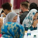 Menschen sitzen dicht an einer langen Tafel in einem Saal beim gemeinsamen Essen; mehrere Frauen tragen Kopftuch.