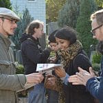 Auf dem Vorhof einer Moschee reicht ein Mann mit Schiebermütze einer lächelnden Frau eine Zeitung; weitere Personen stehen daneben, rechts ein Schild mit Rauchverbot auf Türkisch, Deutsch und Englisch.