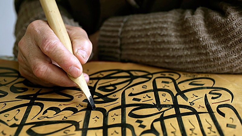 Eine Hand schreibt mit einer Rohrfeder arabische Kalligrafie auf goldfarbenes Papier.