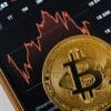 Foto eines physischen Bitcoins auf einer Handy-App, die Crypto-Kurse anzeigt