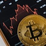 Foto eines physischen Bitcoins auf einer Handy-App, die Crypto-Kurse anzeigt