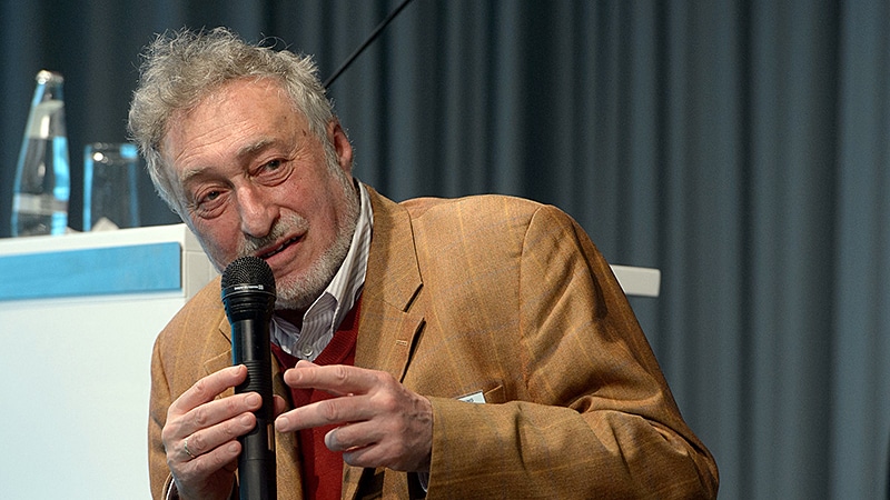 Micha Brumlik, ein älterer Mann mit grauem Bart, spricht auf einem Podium in ein Handmikrofon.