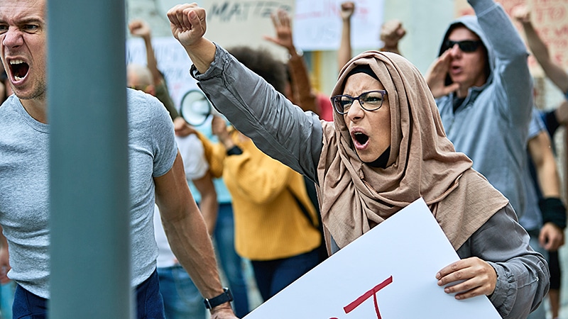 Eine Frau mit Kopftuch reckt bei einer Demonstration die Faust und ruft; sie hält ein Schild, dahinter weitere Demonstranten mit Transparenten.