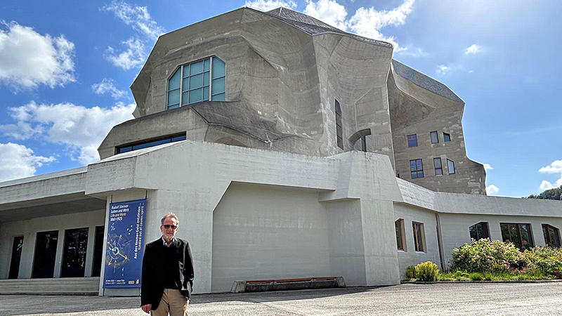 Ein Mann steht vor einem großen, geschwungenen Sichtbetonbau; links am Gebäude hängt ein Plakat zu Rudolf Steiner, „Leben und Werk 1861–1925“.